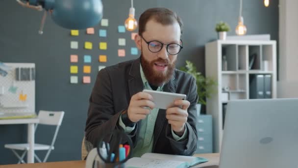 Χαρούμενος τύπος που απολαμβάνει το κινητό βιντεοπαιχνίδι παίζοντας μόνος στο δημιουργικό γραφείο χαλαρώνοντας κατά τη διάρκεια του διαλείμματος — Αρχείο Βίντεο