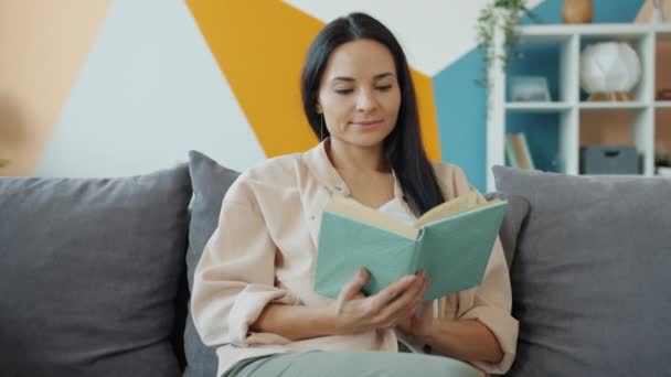 Güzel görünümlü genç bir kadın kitap okuyor ve gülümsüyor. Sonra da güzel bir dairede sayfa değiştiriyor. — Stok video