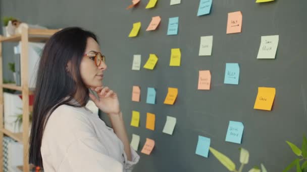 Chica atractiva mirando notas pegajosas brillantes en la pared y pensando en ideas de negocios — Vídeo de stock