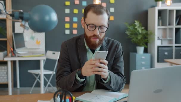 Schöner Geschäftsmann nutzt Smartphone im Büro am Schreibtisch und genießt Online-Kommunikation — Stockvideo