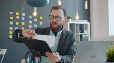 Erkek ofis çalışanı gazete okuyor ve masa başında çalışırken dizüstü bilgisayarını kontrol ediyor.