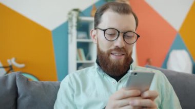 Akıllı telefon mesajları kullanan neşeli genç adam evde yalnız başına eğleniyor ve kanepede dinleniyor.
