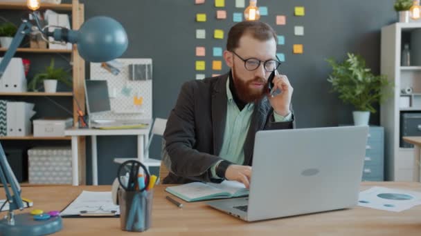 Серйозний хлопець в окулярах офіс працівник пісяє на мобільний телефон працює за столом на робочому місці — стокове відео