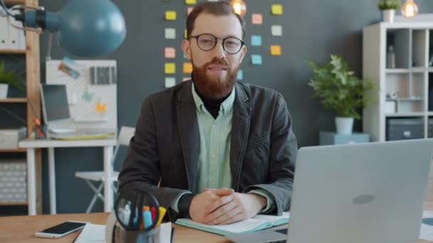 Retrato de jovem empreendedor na mesa no escritório olhando para a câmera com sorriso claro — Vídeo de Stock