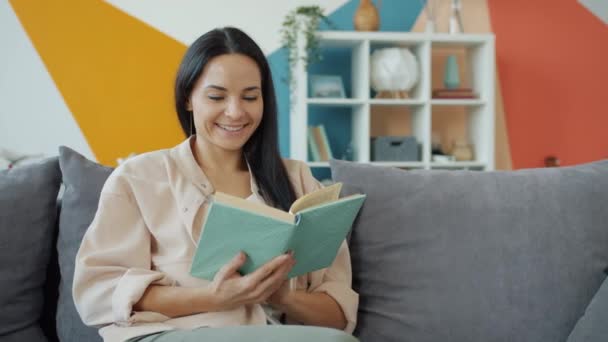 Feliz jovencita disfrutando de libro en casa leyendo y sonriendo relajándose en el sofá solo — Vídeos de Stock