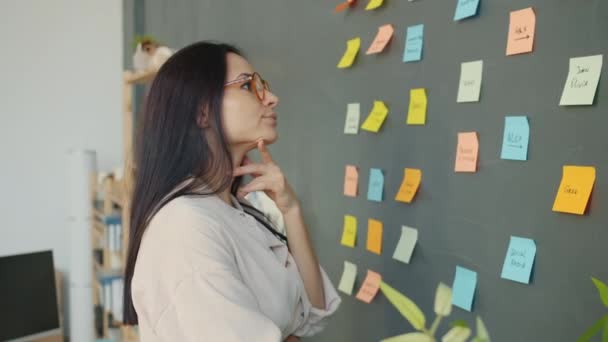 Movimento lento da jovem senhora séria olhando para notas pegajosas na parede do escritório e pensando — Vídeo de Stock