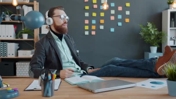 Alegre emprendedor divirtiéndose escuchando música cantando sentado en el escritorio usando auriculares en la oficina — Vídeos de Stock