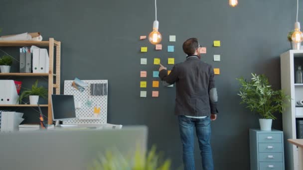 Vista posterior del hombre de negocios leyendo información sobre notas adhesivas en la pared en la oficina creativa — Vídeo de stock