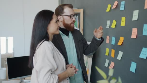 Biznes zespół dziewczyna i facet omawianie pomysłów pracy z lepkich notatek na ścianie biurowej — Wideo stockowe