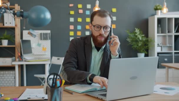 Joven hombre de negocios hablando por teléfono móvil discutiendo problemas de trabajo y el uso de la computadora portátil en la oficina — Vídeo de stock