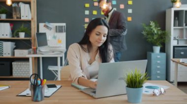 Diz üstü bilgisayar kullanan kadın ofis çalışanı iş yerinde iş belgesini tartışan bir meslektaşla konuşuyor.