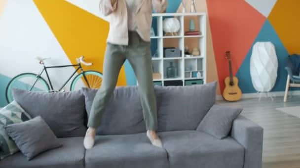 Radosna dziewczyna skacze na kanapie nosząc słuchawki śpiewając w pilocie zabawy — Wideo stockowe