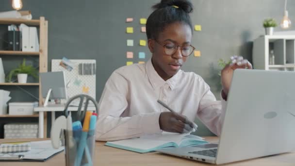 Afro-Amerikaanse zakenvrouw met behulp van laptop het maken van notities in notebook kijken moe en ongelukkig — Stockvideo