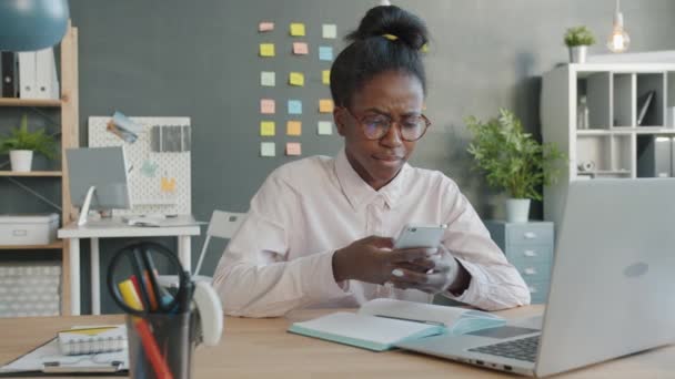 Zwolniony ruch ładnej afro-amerykańskiej dziewczyny korzystającej ze smartfona i uśmiechającej się w miejscu pracy — Wideo stockowe