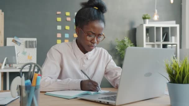 アフリカ系アメリカ人の女の子がノートパソコンを使って職場で不幸な気持ちでノートを書く — ストック動画