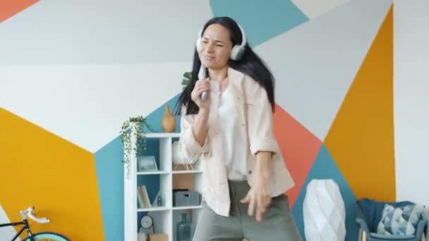 Mujer juguetona bailando en el sofá usando auriculares disfrutando de la música y cantando en control remoto — Vídeos de Stock