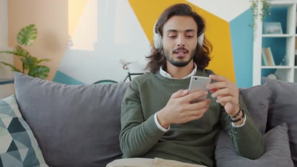 Glad blandras kille i hörlurar sjunga och dansa med smartphone hemma — Stockvideo