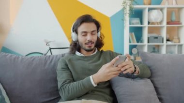 Kulaklık takmış, evde akıllı telefon kullanarak müzik dinleyen neşeli genç bir Arap 'ın ağır çekimleri.