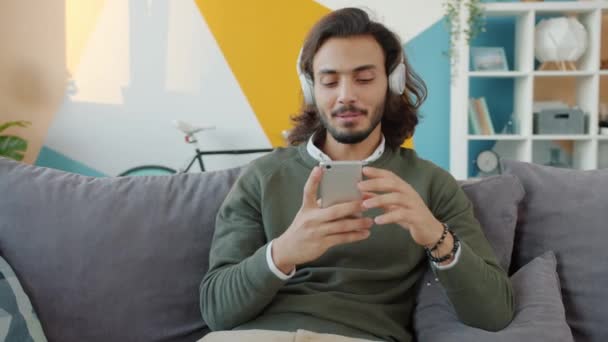 Hombre árabe joven cantando disfrutando de la música en los auriculares y tocando la pantalla del teléfono inteligente en el apartamento — Vídeos de Stock