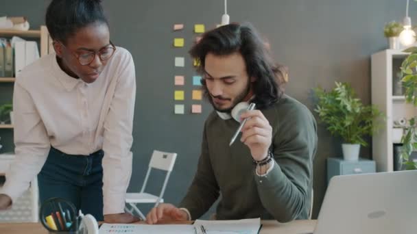 Arabiska killen och afro-amerikanska tjejen tittar igenom pappersarbete och pratar på kontoret — Stockvideo