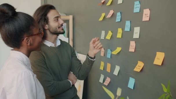 Cheerful coworkers Arab man and Afro-American woman discussing info on sticky notes on office wall — Stock Video