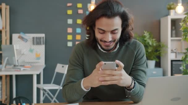 Hombre árabe alegre usando la pantalla táctil del teléfono inteligente y sonriendo en el lugar de trabajo — Vídeo de stock