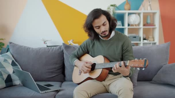 Hombre árabe creativo tocando la guitarra y usando el ordenador portátil viendo tutorial en casa — Vídeos de Stock