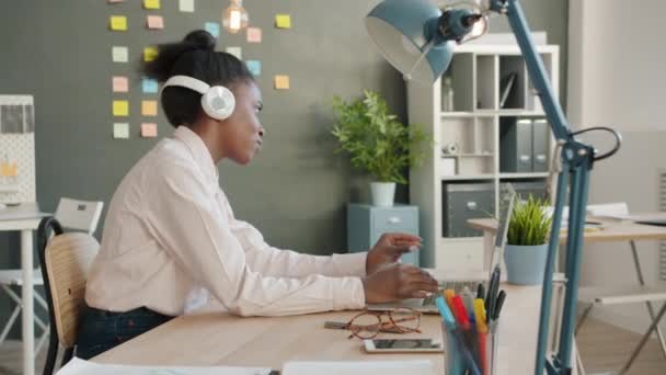 Trabajador de oficina afroamericano relajándose en el trabajo escuchando música y bailando en el escritorio con computadora portátil — Vídeos de Stock