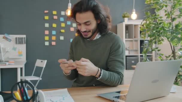 Chico árabe feliz divirtiéndose en el interior de la oficina jugando videojuego móvil en el teléfono inteligente — Vídeo de stock