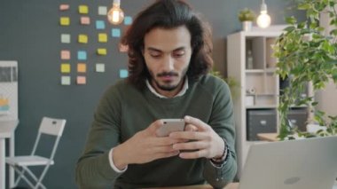 Akıllı telefon kullanan mutlu Arap adam ekranlara dokunuyor ve yaratıcı modern iş yerinde başparmak kaldırıyor.