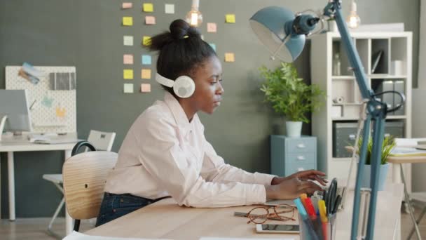 Медленное движение радостной девушки смешанной расы, работающей с ноутбуком, а затем танцующей в наушниках в офисе — стоковое видео