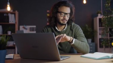 Karma ırk adamı geceleri karanlık ofiste dizüstü bilgisayar kullanarak not defterine bilgi yazıyor.