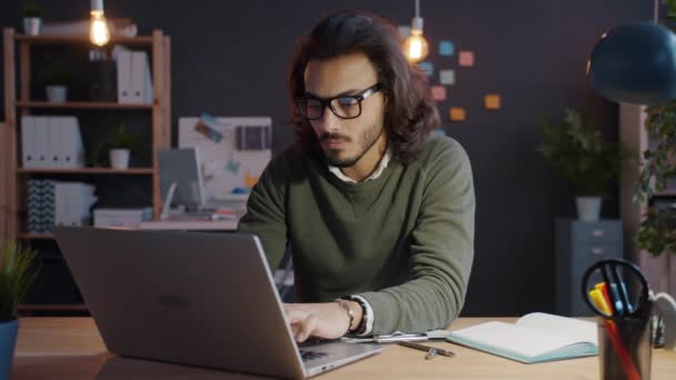 Mouvement lent du gars arabe travaillant avec un ordinateur portable et prenant des notes au bureau la nuit — Video