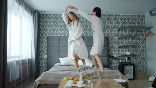 Gadis dan pria dalam mantel mandi menari di tempat tidur di kamar hotel modern bersenang-senang — Stok Video