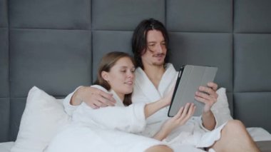 Bornozlu kız ve erkek tablet kullanıyor ekranlara dokunuyor ve otelde yatakta konuşuyorlar.