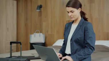 Otel odasında çalışan resmi kıyafetli genç bir kadın dizüstü bilgisayar kullanıyor.