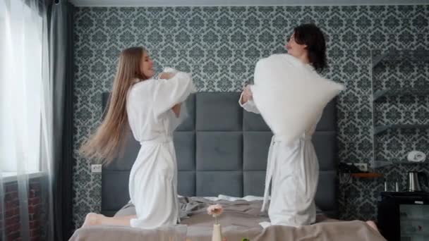 Cámara lenta de pareja alegre en batas de baño luchando almohadas y divirtiéndose en la habitación del hotel — Vídeo de stock