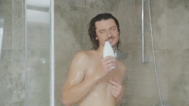 Radosny facet myjący się pod prysznicem i śpiewający w butelce szamponu bawiący się sam — Wideo stockowe