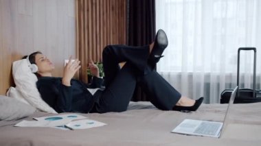 Takım elbiseli mutlu bir kadın otel yatağında kulaklık takıp müzik dinliyor.