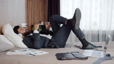 Orta Doğulu takım elbiseli bir adam modern bir otelde yatakta müzik dinliyor.