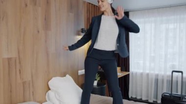 Şık takım elbiseli neşeli bir kızın portresi otel odasında yatakta dans ediyor ve yalnız eğleniyor.
