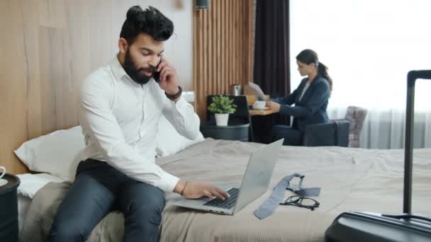 Biznesmen wykonujący telefon komórkowy z pokoju hotelowego i korzystający z laptopa podczas pracy kobiety w tle — Wideo stockowe