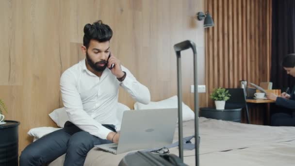 Facet rozmawiający przez telefon komórkowy i używający laptopa w pokoju hotelowym, kobieta pracująca w tle — Wideo stockowe