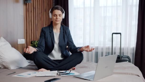 Ung affärskvinna i hörlurar mediterar i lotusförvaring på hotellsäng — Stockvideo