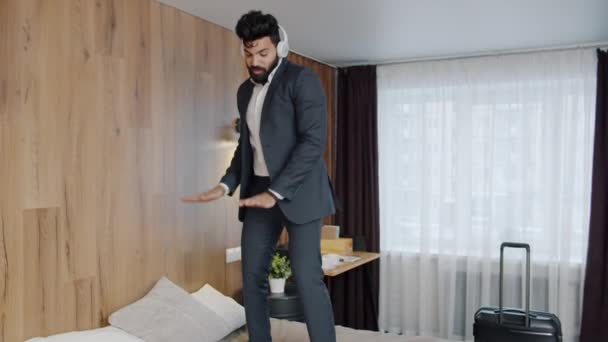 Happy Bliski Wschód człowiek noszenia słuchawki taniec na łóżku w pokoju hotelowym — Wideo stockowe