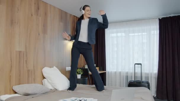Szczęśliwa młoda kobieta w garniturze tańczy na łóżku hotelowym ciesząc się wakacjami nosząc słuchawki — Wideo stockowe