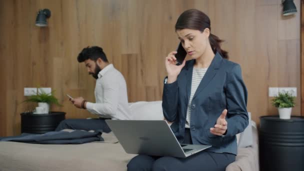 Bizneswoman rozmawia przez telefon komórkowy pracując z laptopem w pokoju hotelowym podczas gdy facet używa smartfona — Wideo stockowe