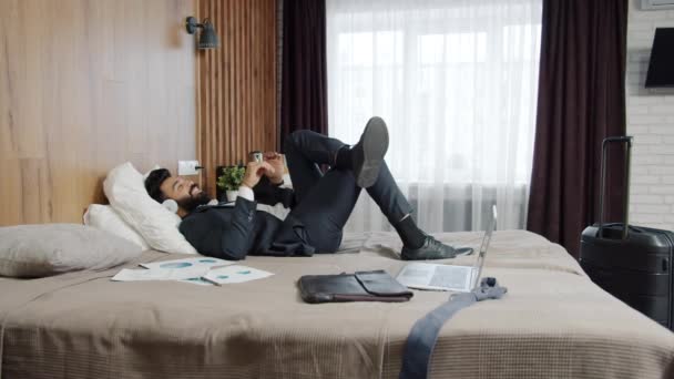 Szczęśliwy arabski biznesmen korzystających z muzyki poprzez słuchawki relaks w łóżku hotelowym — Wideo stockowe