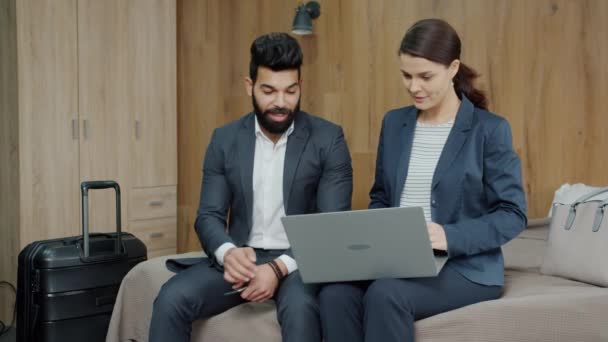 Νέοι συνεργάτες που χρησιμοποιούν φορητό υπολογιστή και μιλούν καθισμένοι στο κρεβάτι στο δωμάτιο του ξενοδοχείου — Αρχείο Βίντεο