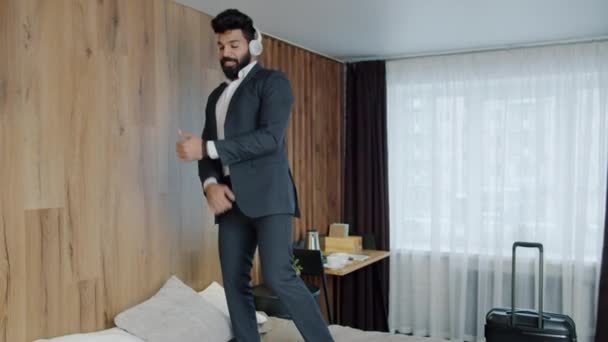 Радостный арабский бизнесмен танцует на кровати отеля, наслаждаясь музыкой через наушники — стоковое видео